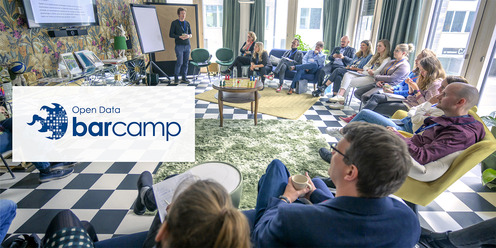 Mehrere Personen sitzen in einem bunten Raum im Halbkreis. Vorne steht ein Moderator neben einem Monitor. Links im Bild ist das Open Data Barcamp Logo
